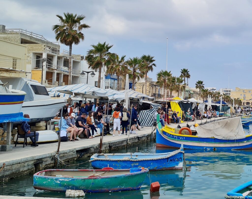 Blick auf die Promenade, auf der viele Menschen sitzen. Dahinter Stände mit Marktschirmen, Palmen und Häuser. Im Wasser drei blaue Boote.