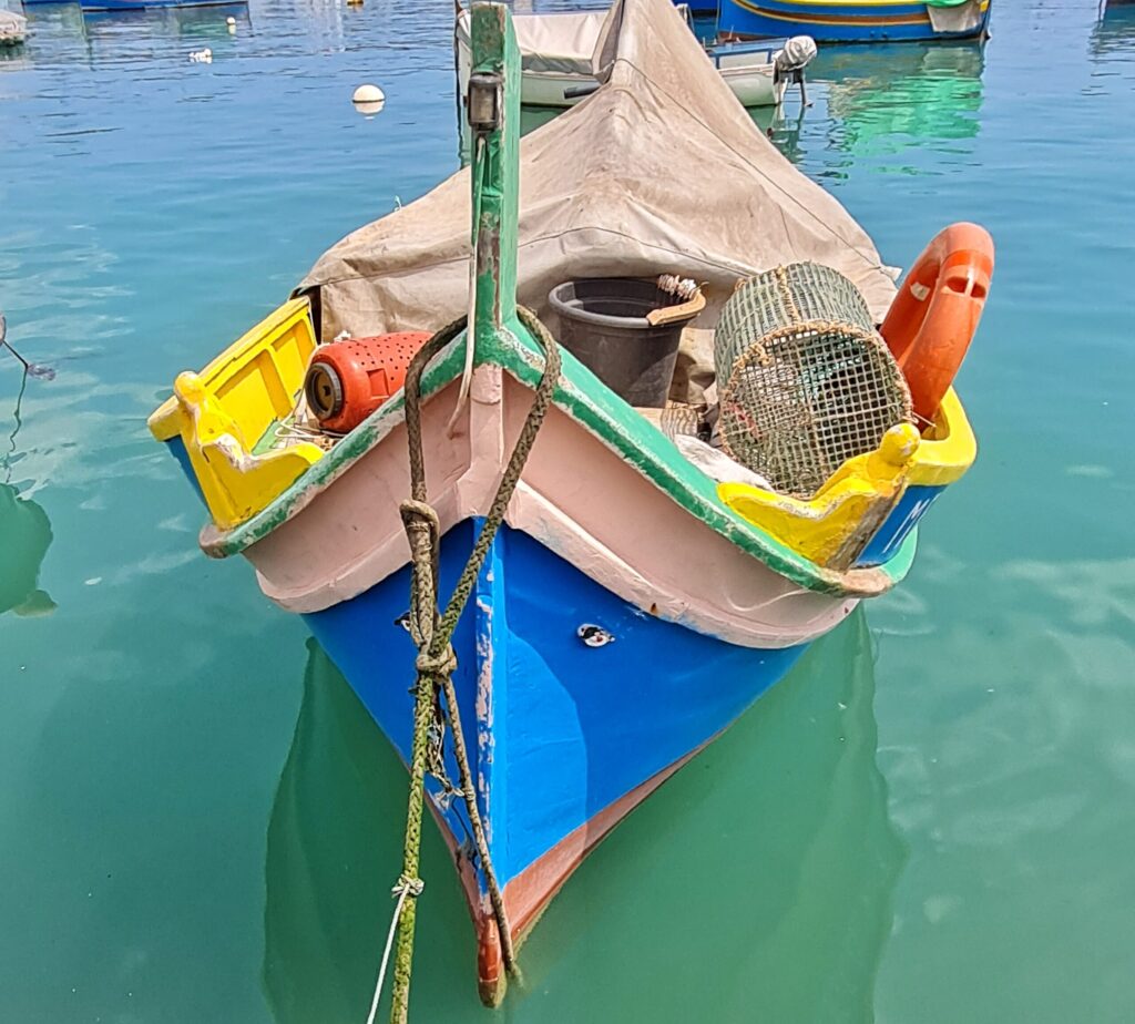 Ein Boot , das im Wasser liegt, aus der Nähe fotografiert. Der untere Teil ist blau und darüber ein blasses Rosa. Der innere Teil des Bootes ist weitgehend von einer Plane, einem Eimer, einer Reuse, einem orangenen Rettungsring und einem orangenen Behälter verdeckt. Man erkennt aber, dass das Innere des Bootes in einem leuchtenden Gelb gestrichen ist. Vone sind auf dem blauen Teil rechts und links zwei kleine Augen angebracht.