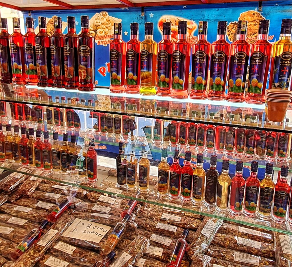 Ein Stand mit mehreren Reihen von Flaschen, die Flüssigkeiten in Rot oder Gelb. Auf den Etiketten sind Früchte zu sehen. Im unteren Bereich durchsichtige Beutel, die Nüsse oder ähnliches enthalten. Darauf liegt ein Zettel: "1 = 3 Euro. 3 = 20 Euro".