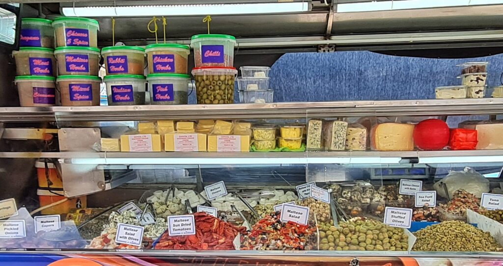 Eine Auslage. Ganz unten mit Oliven, getrockneten Tomaten, Fischsalate und weißem Käse. Darüber eine Reihe von unterschiedlichen Käsestücken. Darüber Plastikbehälter mit Gewürzen und Oliven.