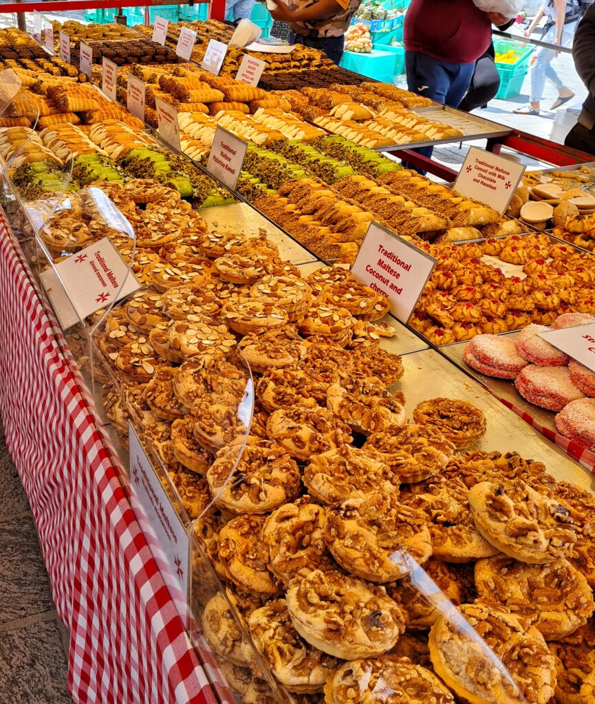 Ein Stand mit verschiedenem Gebäck. Die Sorten sind jeweils mit kleinen Schildern gekennzeichnet, zum Beispiel "Traditional Maltese Coconut and Cherry".
