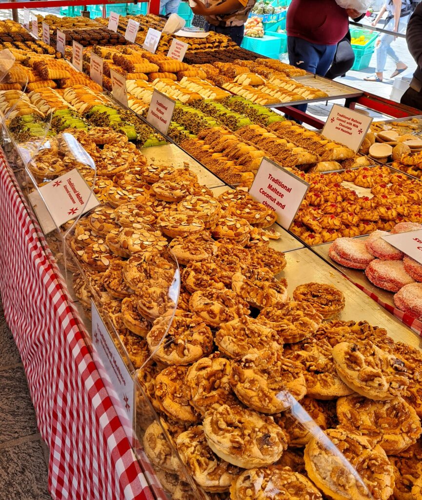 Ein Stand mit verschiedenem Gebäck. Die Sorten sind jeweils mit kleinen Schildern gekennzeichnet, zum Beispiel "Traditional Maltese Coconut and Cherry".