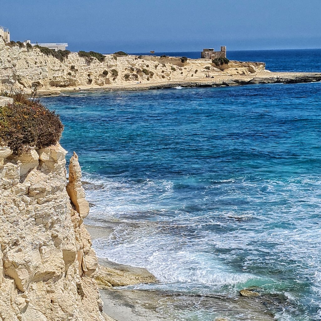 Im oberen Teil eine felsige Landzunge, an deren Spitze ein kleines festungsartiges Gebäude steht. Links ebenfalls Felsen. Im übrigen blaues Meer, teilweise mit weißer Gischt.