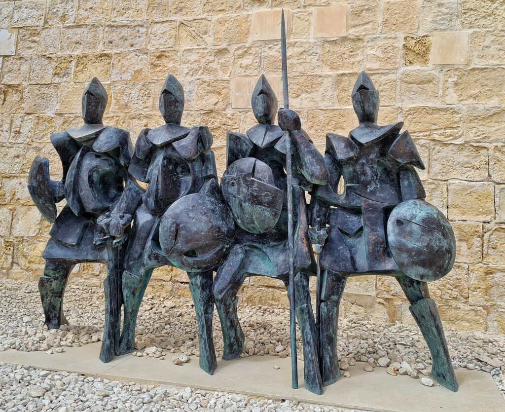 Vor einer gelblich-weißen Ziegelwand stehen vier Figuren aus Metall dicht nebeneinander. Sie tragen Helme und Visiere, Rüstungen, Schilde, Schwerter und Stöcke.
