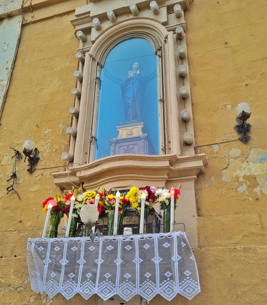 Eine Nische in einer Hauswand in dunkelgelber Farbe. Die Nische ist verglast. Darin steht auf einem sargähnlichen Podest eine Frauenstatue in einem langen Gewand mit einem blauen Umfang. Sie hat die Hände gefaltet und trägt einen Heiligenschein. Um die Nische herum sind kleine Lampen angebracht. Sie sind ausgeschaltet. Unter der Nische ist eine Art Balkon mit Gittern, aber sehr klein. Darauf stehen mehrere Vasen mit Blumen, ausserdem Kerzen mit LED-Licht. Sie sind eingeschaltet. An dem Balkon hängt unten eine niedrige, weiße Gardine.