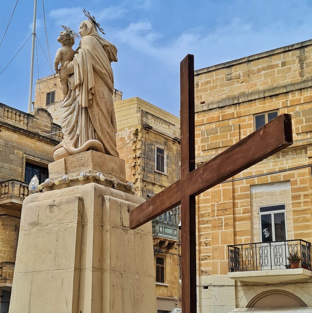 Inmitten von gelblichen Häusern eine Steinstatue auf einem hohen Sockel. Sie zeigt Maria mit dem Jesuskind auf dem Arm. Die Aufnahme zeigt sie leicht seitlich von hinten. Im Vordergrund ein großes Holzkreuz.