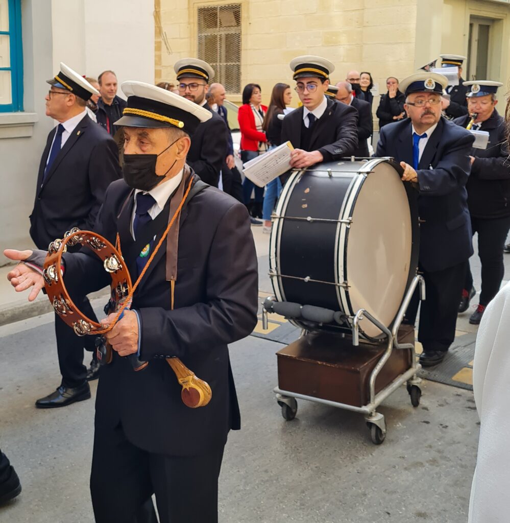 Ein Teil einer Musikkapelle, bestehend aus mehreren Männern unterschiedlichen Alters. Sie tragen dunkelblaue Anzüge mit blauen Krawatten und weiße Mützen mit dunklen Schirmen mit einer Goldkordel. Der vorderste Mann spielt ein Tamburin. Hinter ihm ein Mann, der auf einem kleinen Rollwagen eine große Trommel schiebt. Die Männer im Hintergrund spielen verschiedene Blasinstrumente.
