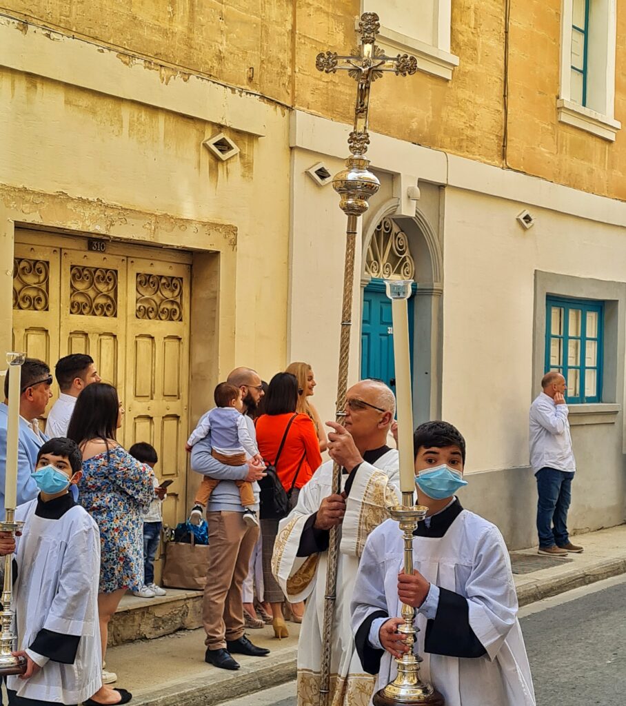 Ein Teil einer Gasse mit gelblich-beigen Häusern und farbigen Türen und Fenstern. Auf dem schmalen Gehsteig stehen einige Menschen, teilweise mit Kindern und schauen nach etwas, das auf dem Fotoausschnitt nicht zu sehen ist. Auf der Straße zwei Meßdiener mit hellblauen medizinischen Masken. Sie tragen jeweils einen großen silbernen Leuchter mit hohen LED-Kerzen. Hinter ihnen ein Priester in einem beigen Gewand, teilweise golden bestickt. Er trägt einen hohen silbernen Stab, auf dem oben ein Kreuz ist.