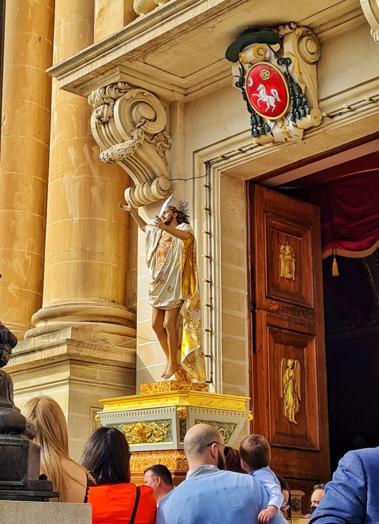 Ein geöffnetes Kirchenportal. Über dem Portal ein rotes Wappen mit einem weißen Pferd darin. Herausgetragen wird eine große Christusfigur mit erhobenen Armen. Sie steht auf einem sargähnlichen, weiß-goldenen Podest. Christus hat ein langes Tuch übergeworfen, das außen weiß mit goldenen Mustern ist und innen goldfarben. Man sieht die Hinterköpfe der Menschen, die davor stehen. Ein Mann hat sein Kind auf den Arm genommen.