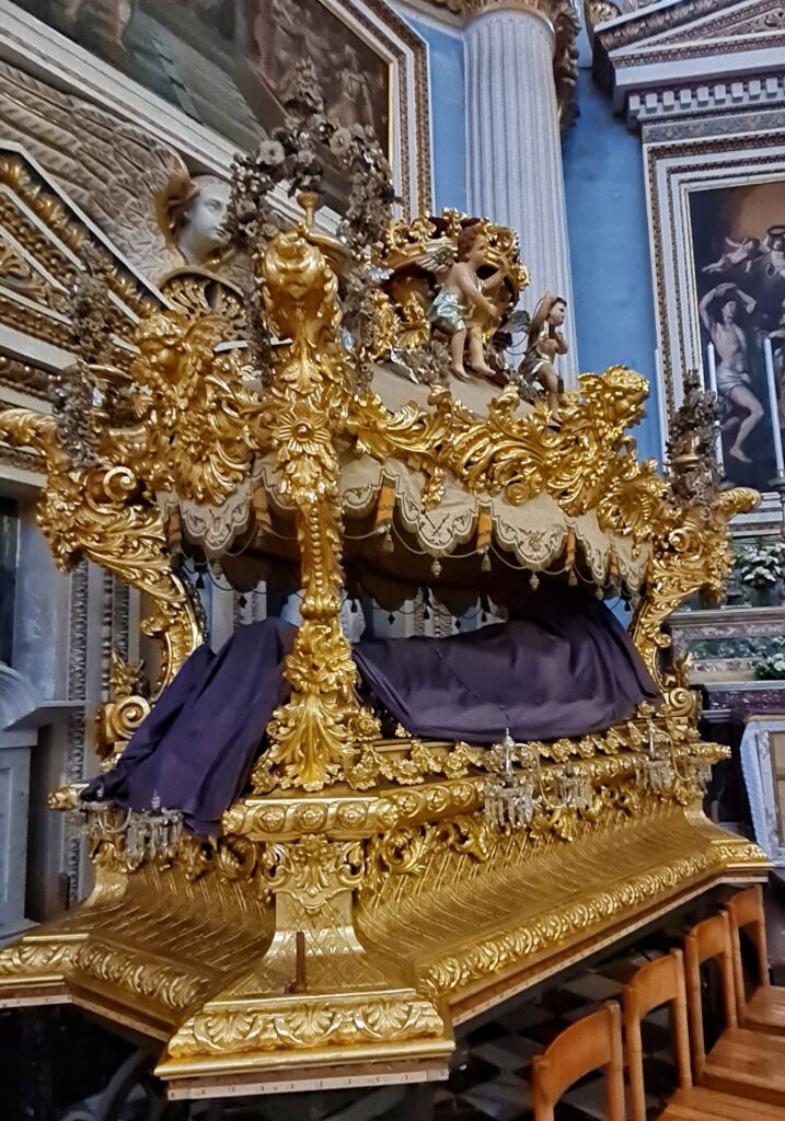 In einem Raum mit prächtigen Gemälden, Engelsfiguren, Säulen und goldenen Verzierungen an den Wänden steht etwas, das aussieht wie ein geöffneter goldener Sarg, wobei zwischen dem unteren Teil und dem Sargdeckel an den Ecken kleine Pfosten angebracht sind. Dazwischen liegt eine Figur, die vollständig von einem auberginefarbigen Tuch bedeckt ist.