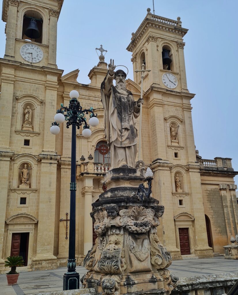 Eine Kirche aus hellem Stein von vorne fotografiert. Sie hat zwei eckige Türme. Im oberen Teil sind durch die Öffnungen kleine Glocken zu erkennen. Darunter jeweils eine runde Scheibe. Die rechte zeigt die Uhrzeit, auf der linken sind aussen Zahlen und innen Worte mit drei Buchstaben zu erkennen. Darunter jeweils zwei Nischen mit Steinfiguren darin. Ganz unten jeweils eine Holztür mit Kassettenverzierungen. Die linke ist halb geöffnet. Die Mitte der Kirche ist weitgehend verdeckt durch eine hohe Steinstatue auf einem großen verzierten Steinpodest. Sie zeigt einen alten Mann in einem langen Gewand mit Falten und einem breiten herabhängenden Schal. Er hat einen Heiligenschein. In seiner linken Hand (aus Sicht der Kamera also rechts) hat er einen hohen Stab, oben mit einem Kreuz. Die andere Hand hat er segnend erhoben.