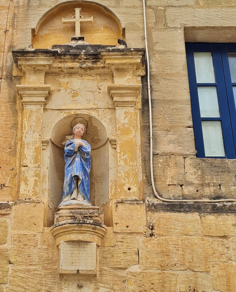 Ein Teil einer Hauswand aus gelblichem Stein. In einer Nische steht eine Frauenfigur aus Stein in einem blauen Gewand mit einem Heiligenschein. Sie ist etwas verwittert oder schmutzig. Die Nische ist verziert mit einem Portal. Darüber ist ein Kreuz. Rechts ist ein Teil eines Fensters mit dunkelblauen Sprossen zu sehen.