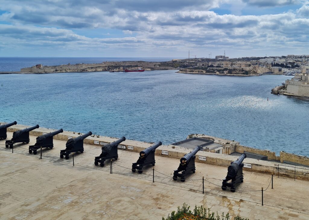 Blick von oben auf eine große Terrasse, auf der nebeneinander Kanonen ausgerichtet sind. Sie "zielen" auf eine große Wassefläche und auf das gegenüberliegende, flache Ufer, das mit Gebäuden bebaut ist.