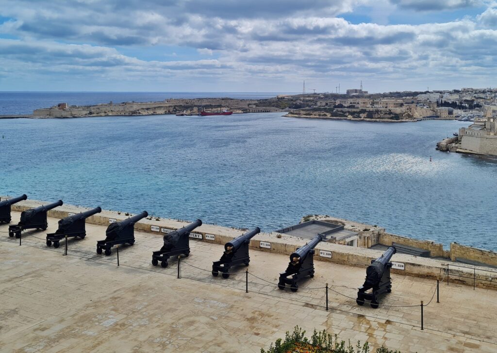 Blick von oben auf eine große  Terrasse, auf der nebeneinander Kanonen ausgerichtet sind. Sie "zielen" auf eine große Wassefläche und auf das gegenüberliegende, flache Ufer, das mit Gebäuden bebaut ist.