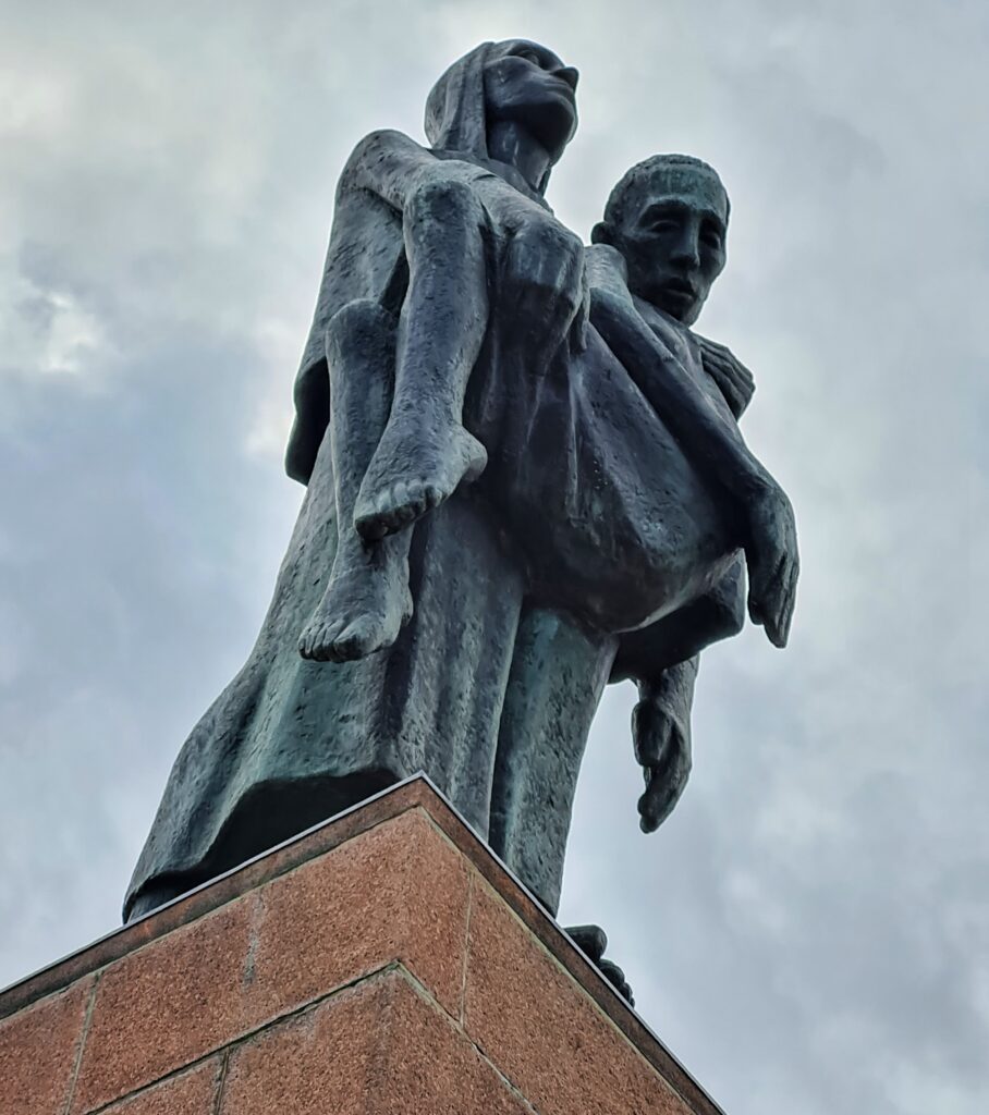 Ein Denkmal von seitlich-unten aufgenommen. Es zeigt eine aufrecht stehende Person, die in ihren Armen eine andere Person trägt. Deren Arme und Beine hängen nach unten.