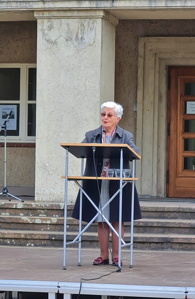 Eine kleine, alte Frau mit weißen Haaren steht an einem Redepult. Hinter ihr ein paar Stufen zu einem Gebäude, von dem ein breiter Pfeiler und die linke Seite einer großen Holztür zu erkennen ist.