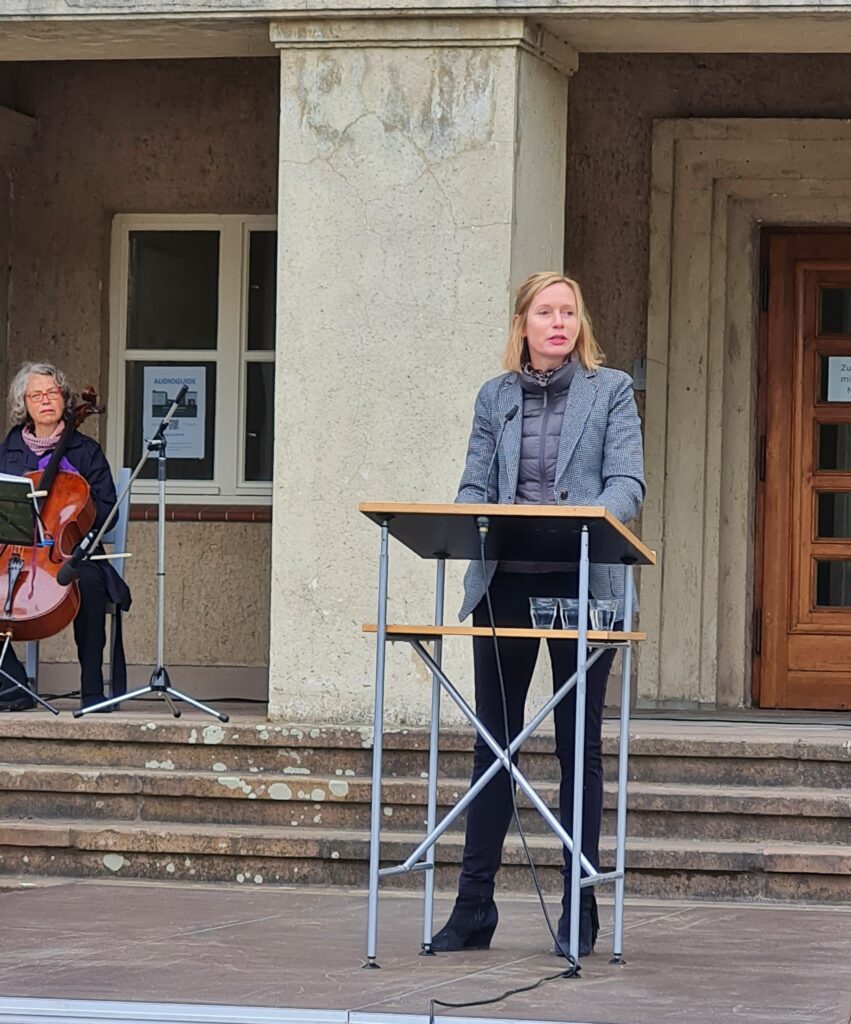 Eine junge Frau mit halblangen, blonden Haaren steht an einem Redepult. Sie trägt eine dunkle Hose und ein graues Jackett, darunter eine graue Steppweste. Hinter ihr ein paar Stufen zu einem Gebäude mit einem breiten Pfeiler. Links davon eine ältere, auf einem Stuhl sitzende Frau in dicker Jacke mit einem großen Streichinstrument. Rechts vom Pfeiler ist ein Teil einer großen Holztür zu sehen.