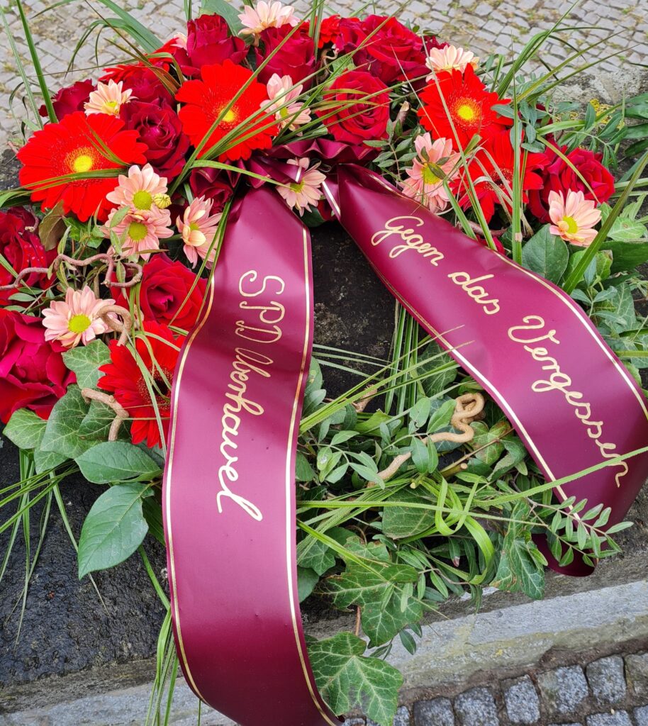 Ein Kranz mit Blumen in verschiedenen Rottönen. Daran eine Kranzschleife mit der Aufschrift "Gegen das Vergessen SPD Oberhavel".