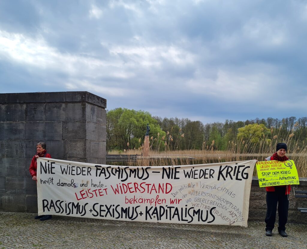 Links eine hohe, graue Mauer, die rechts deutlich niedriger wird, so dass man im Hintergrund Bäume, eine Statue auf einer hohen viereckigen Säule und Schilf sieht. Im Vordergrund zwei Frauen, die zwischen sich ein großes, weisses Transparent halten. Der Text darauf lautet:"Nie wieder Faschismus - nie wieder Krieg heißt damals und heute leistet Widerstand bekämpfen wir Rassismus + Sexismus + Kapitalismus. Das politische Vermächtnis der Widerstanskämpferinnen und Überlebenden aktiv weitertragen".Die rechte Frau hat ausserdem ein gelbes Schild. Von dem Zext darauf sind nur die in Großbuchstaben geschriebenen Worte deutlich zu erkennen:"Frauenwiderstand gegen Kriege Stop dem Krieg Nein Aufrüstung Kriegstreiberei EU NATO USA"