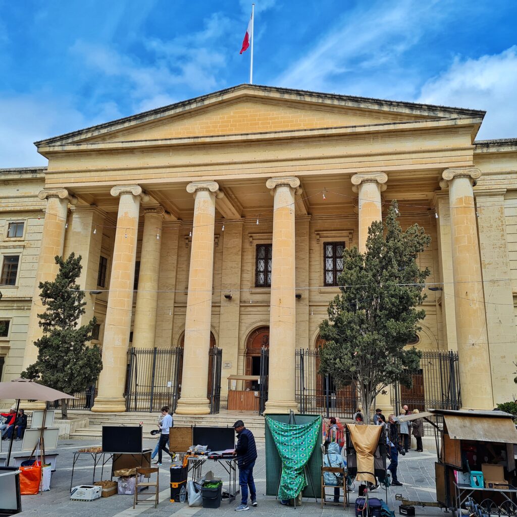 Ein Gebäude mit hohen Säulen im Eingangsbereich. Zwische den Säulen ist unten ein Zaun. In der Mitte ist eine Öffnung, hinter der ein Schalter zu sehen ist. Zu dem Gebäude führen einige breite Treppenstufen. Auf dem Platz davor einige Straßenstände.