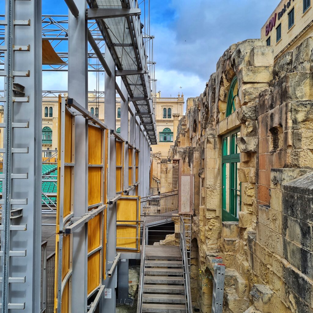 Rechts eine alte, freistehende Mauer mit Fenstern. Links ein Metallgerüst, teilweise gelb verkleidet. In der Mitte geht (ungefähr bis zur Mitte des Fotos) eine schmale Metalltreppe hoch.