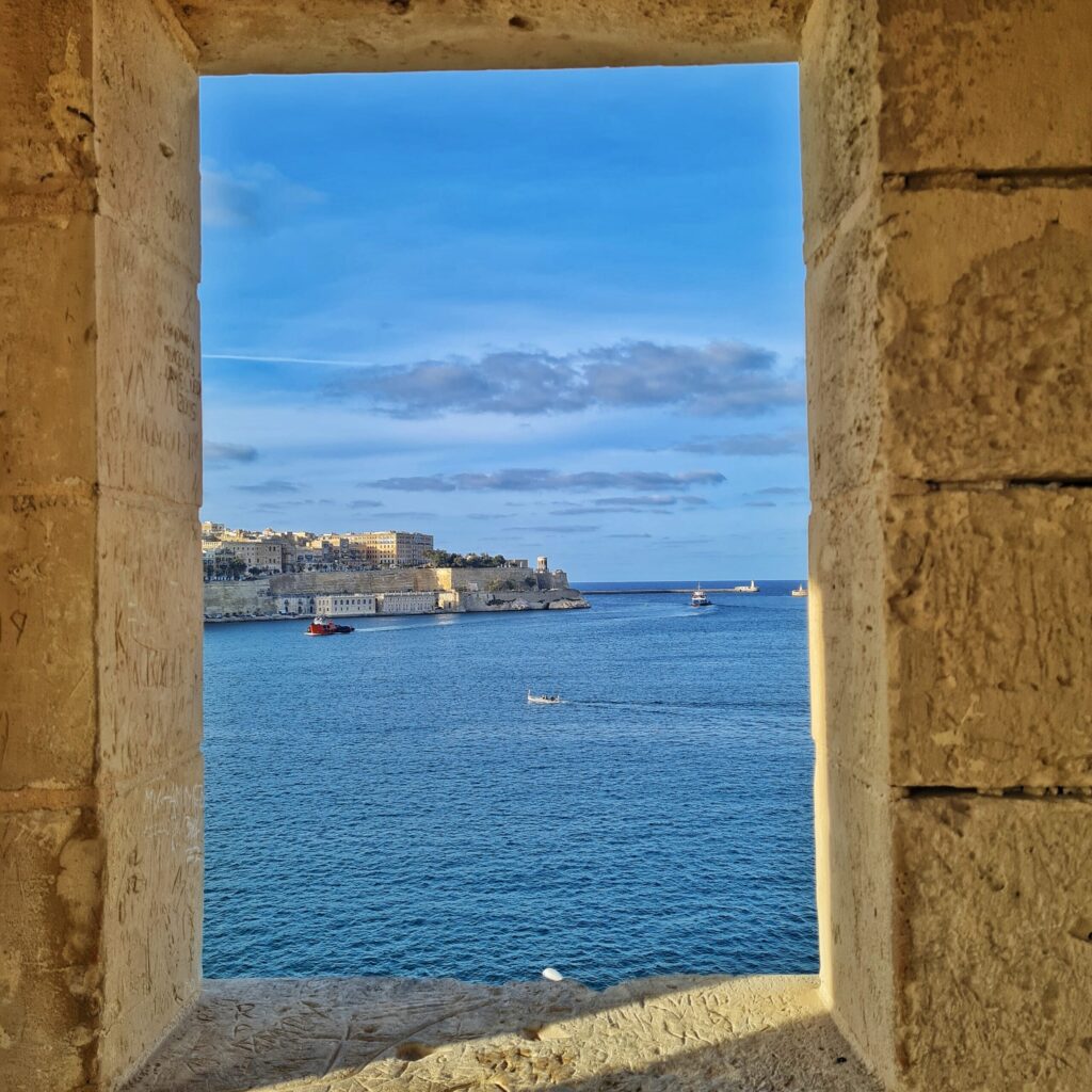 Durch eine rechteckige Öffnung  in einer Steinmauer blickt man auf Wasser, auf dem im Hintergrund einige Boote fahren, und eine Landzunge, bebaut mit viereckigen gelblichen Gebäuden.
