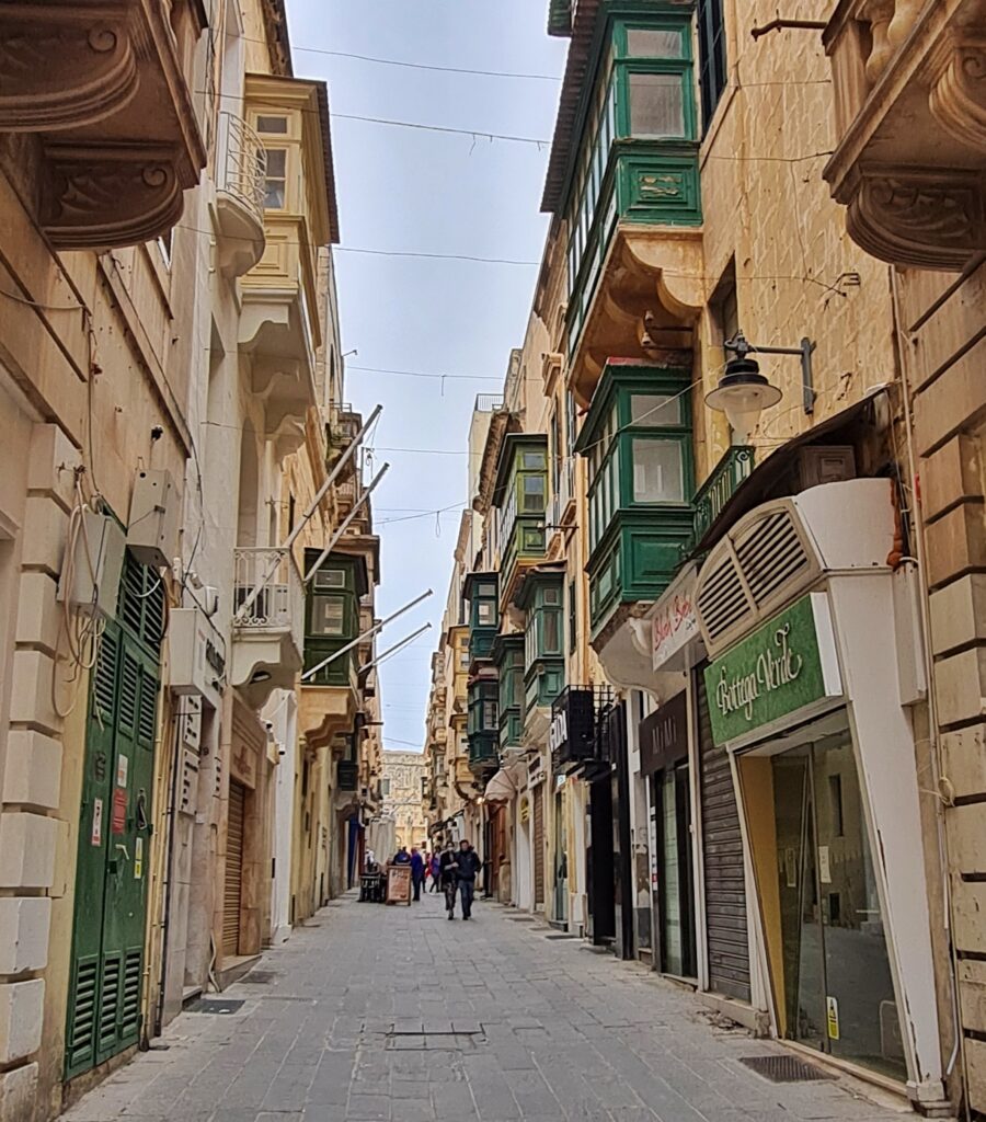 Eine schmale Gasse. Links und rechts durchgehende dreistöckige Häuserreihen. Die Häuser haben maltesische Balkone, die meisten in dunkelgrün. Im Erdgeschoß sind kleine Läden und geschlossene Garagentore oder Rolläden. Im Hontergrund laufen einige Menschen