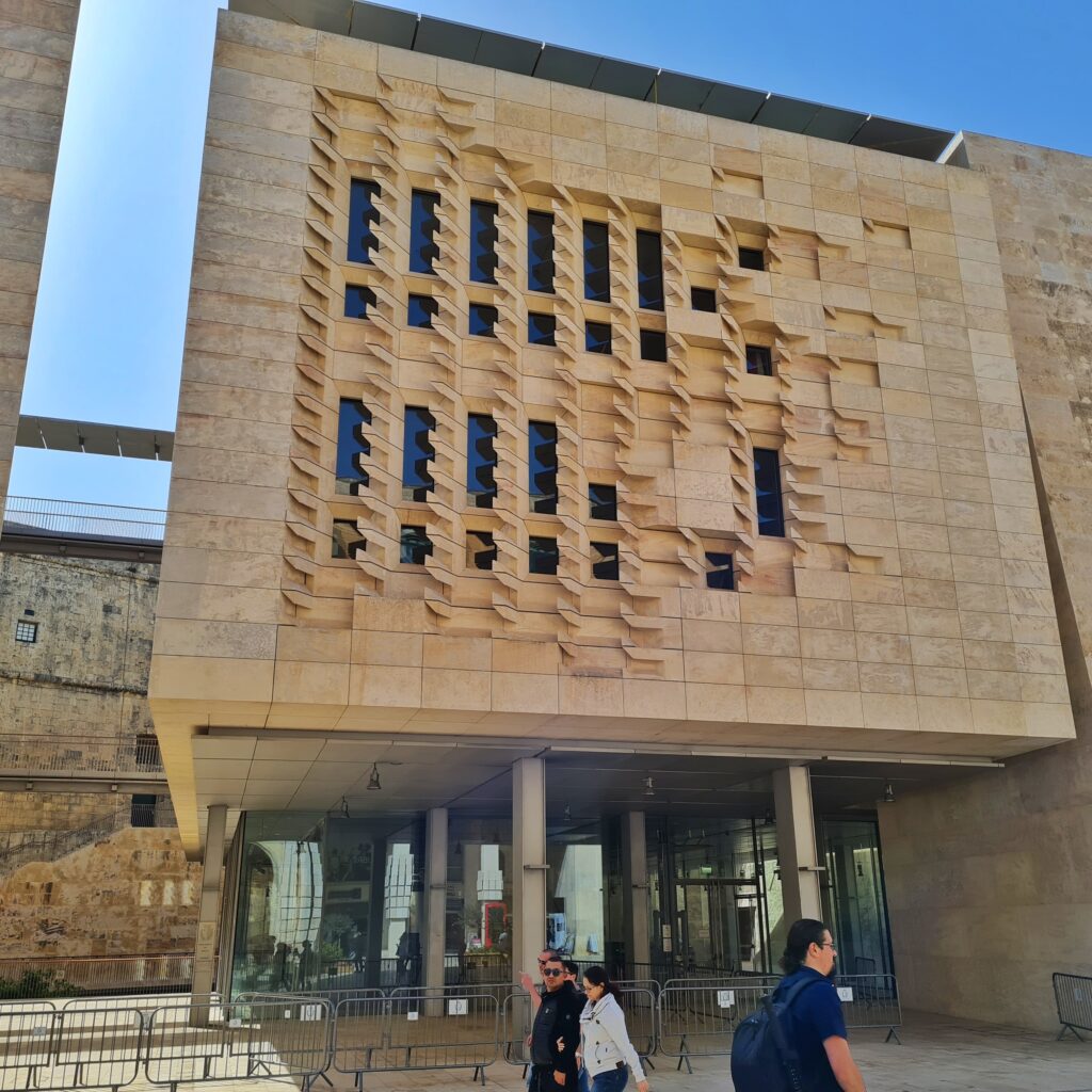 Ein quadratischer Gebäudeteil mit schmalen Fenstern, die ein Muster bilden. Er ruht auf schmalen Säulen; dahinter ist eine Glasfront.