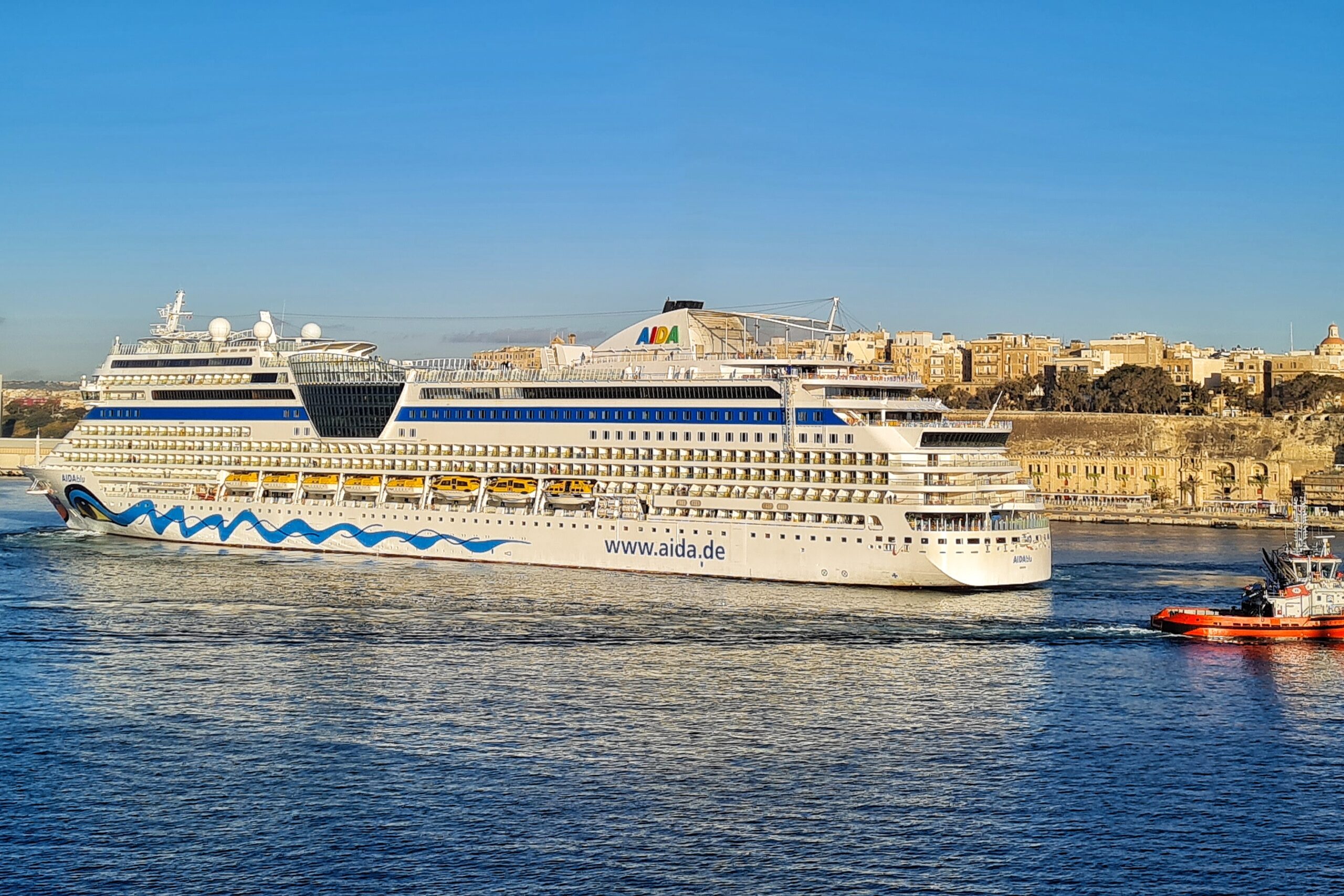 Ein weißes Kreuzfahrtschiff mit der Aufschrift "www.aida.de" fährt entang des Kais von Valletta. lDahinter ein roter Schlepper.