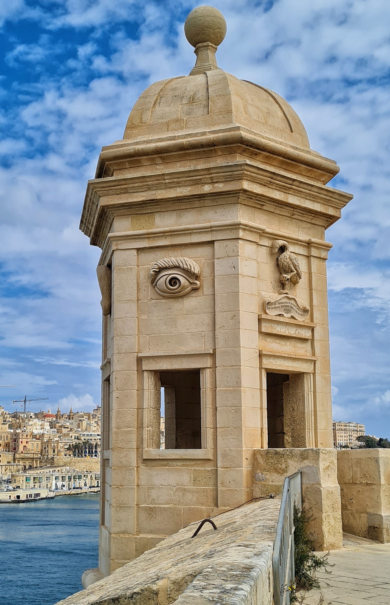 Der Turm, etwas mehr entfernt. Der Vogel ist jetzt auf der linken Seite, und an der anderen Turmwand ist ein Auge. Linksdavon sieht man das gegenüberliegende Ufer mit dichter Bebauung.