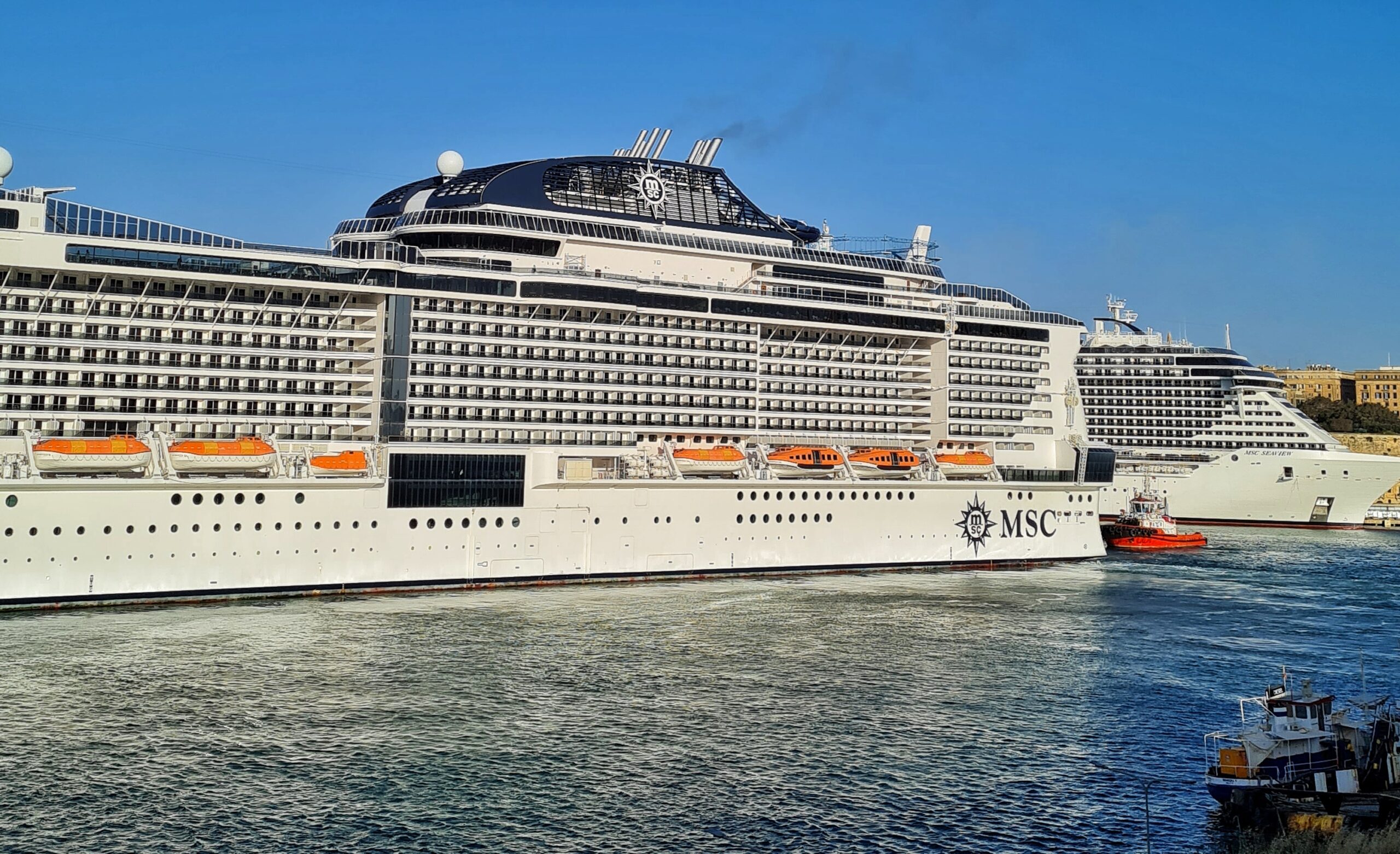 Ein Teil eines hohen weißen Kreuzfahrtschiffes mit der Aufschrift "MSC". Dahinter ein roter Schlepper und quer dazu ein Teil eines weiteren Kreuzfahrtschiffes.