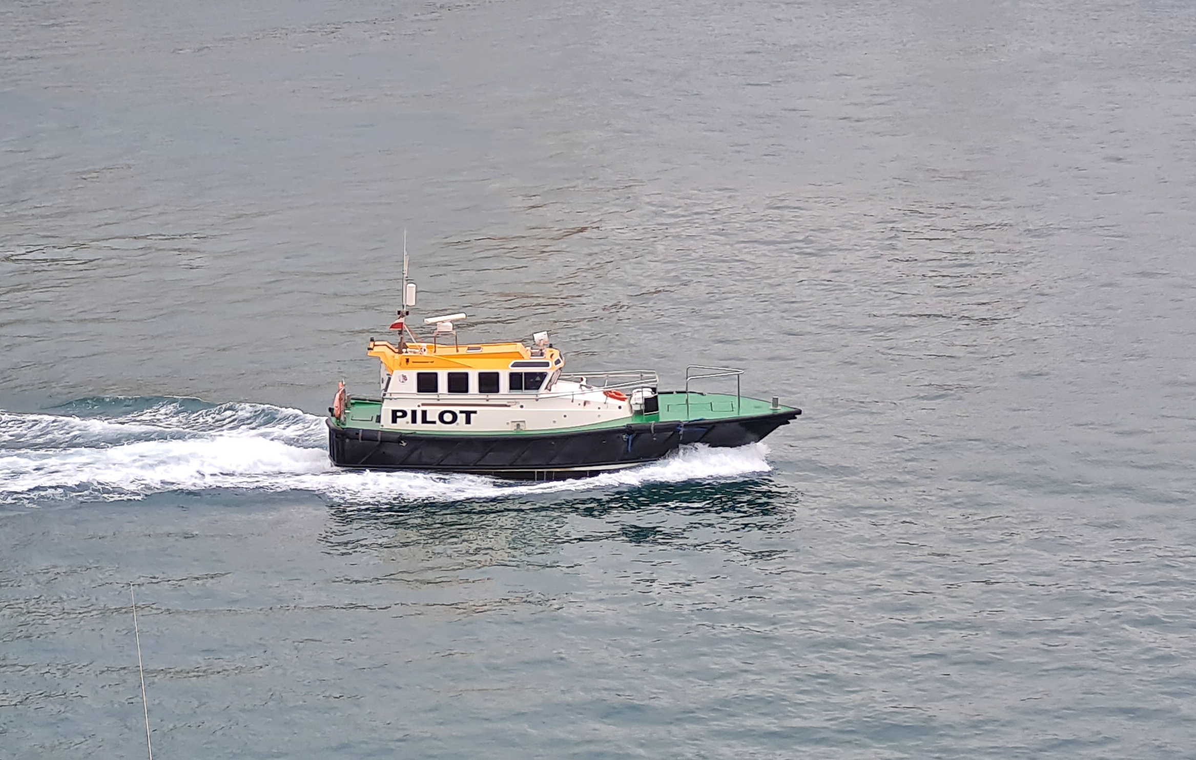 Ein kleines Schiff. Der Rumpf ist schwarz, das Deck grün und das Dach der Kajüte hellorange. Auf dem Schiff steht "Pilot".