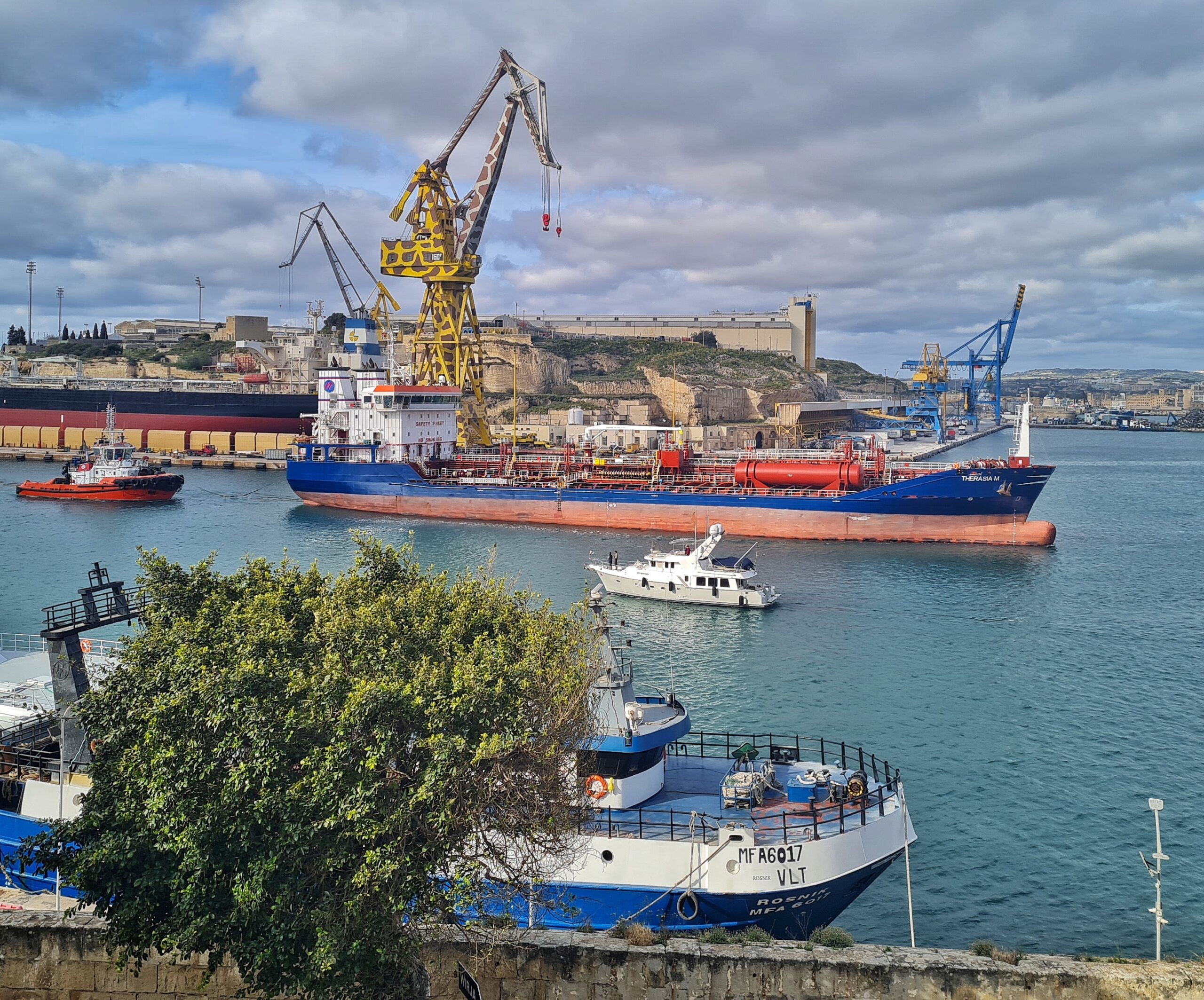 Vor dem Dock, in dem das große Schiff neben dem "Giraffen-Kran" liegt fährt ein großes Frachtschiff nach rechts, dahinter ein roter Schlepper. Nach links fährt eine kleine weiße Yacht. Am unteren Ufer liegt ein Schiff,  größtenteils durch einen  Baum verdeckt.