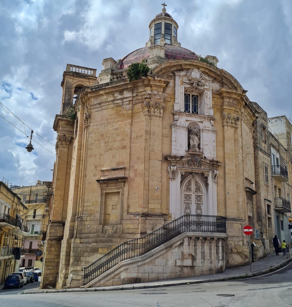 Eine kleine Kirche direkt an einer Straße. Zum Eingang führt eine Treppe mit Geländer hoch. Über der Eingangstür eine verwitterte weiße Statue. Darüber das einzige erkennbare Fenster. Oben eine kleine Kuppel. Darüber ein Türmchen mit Glas und einem Kreuz darauf. Im Dachbereich wachsen Pflanzen.