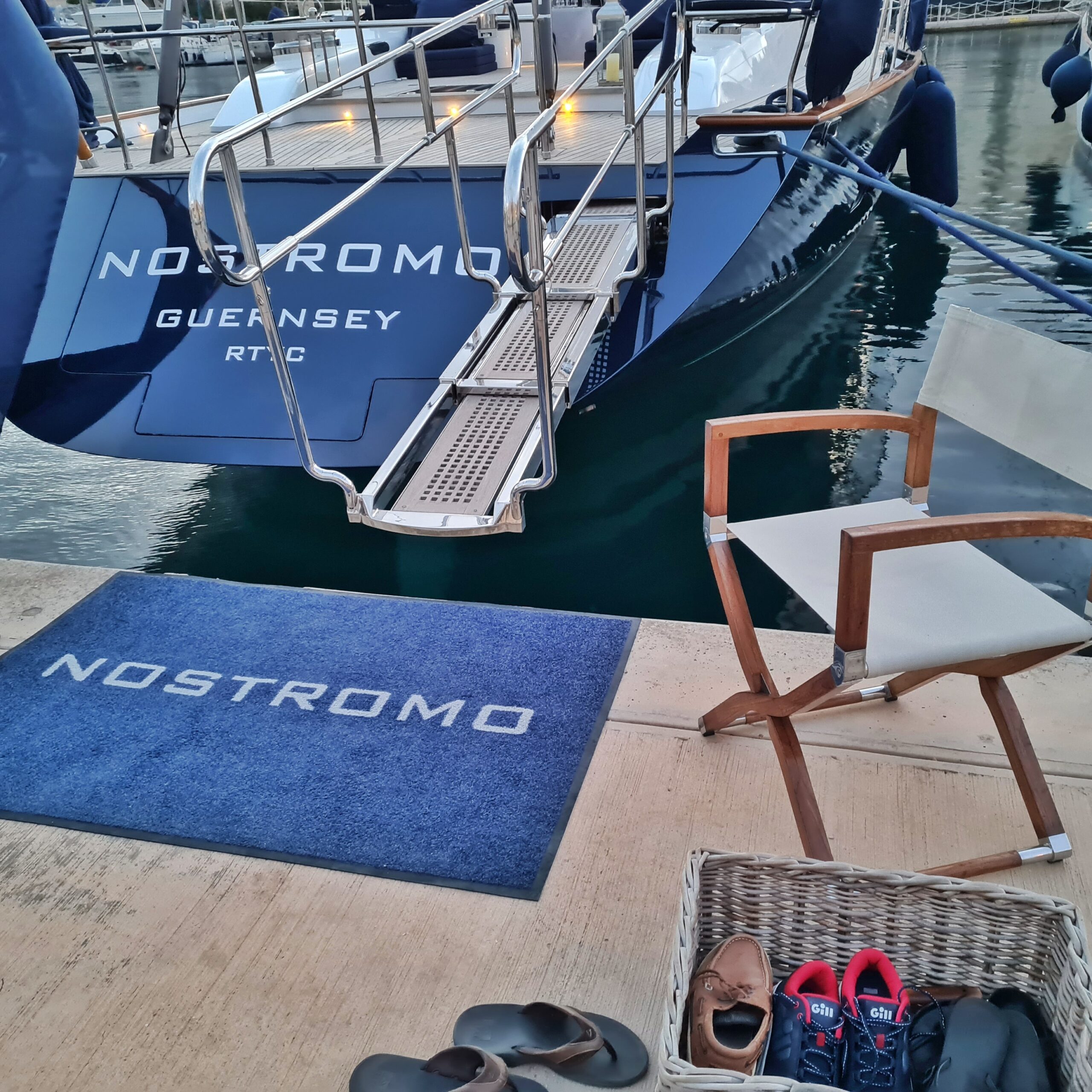 Das Heck einer im Wasser liegenden blauen Yacht. Der angebrachte Name ist nicht vollständig zu erkennen; man sieht nur die Buchstaben "OSTROMO", darunter "Guernsey". Auf dem Kai liegt eine große blaue Fußmatte, auf der ebenfalls die Buchstaben "OSTROMO" zu erkennen sind. Rechts davon steht ein Holzklappstuhl mit weißem Stoff und davor ein viereckiger Korb mit Sneakern und Halbschuhen. Daneben steht ein Paar Pantoletten.