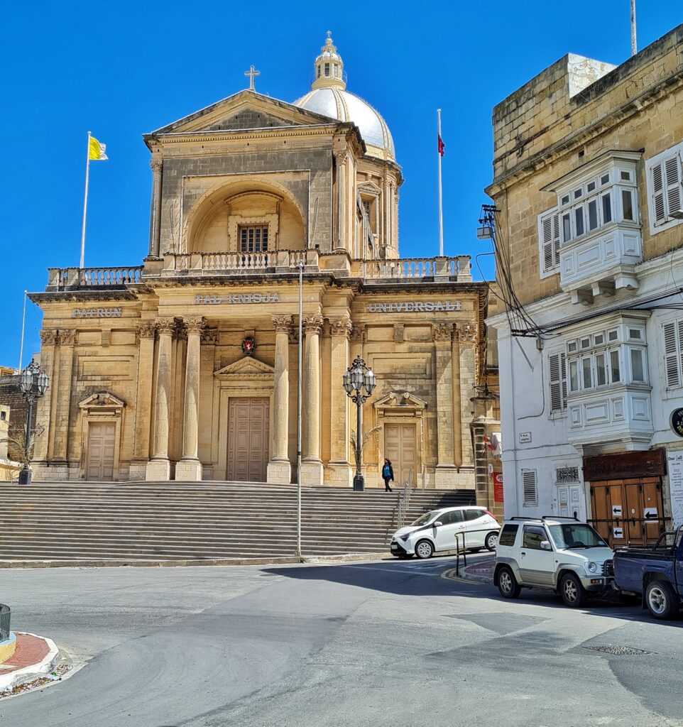 Eine beige Kirche an einer Straße. Rechts ein weiteres Gebäude und parkende Autos. Zur Kirche führen mehrere niedrige Treppenstufen empor.  Die drei Türen (die mittlere mit Säulen davor) sind geschlossen. Darüber eine Dachterrasse mit einem Aufbau in der Mitte. Dort weht eine gelb-weiße Flagge. Im Hintergrund ein runder Turm mit einem kuppelförmigen Dach und einem Türmchen darauf.