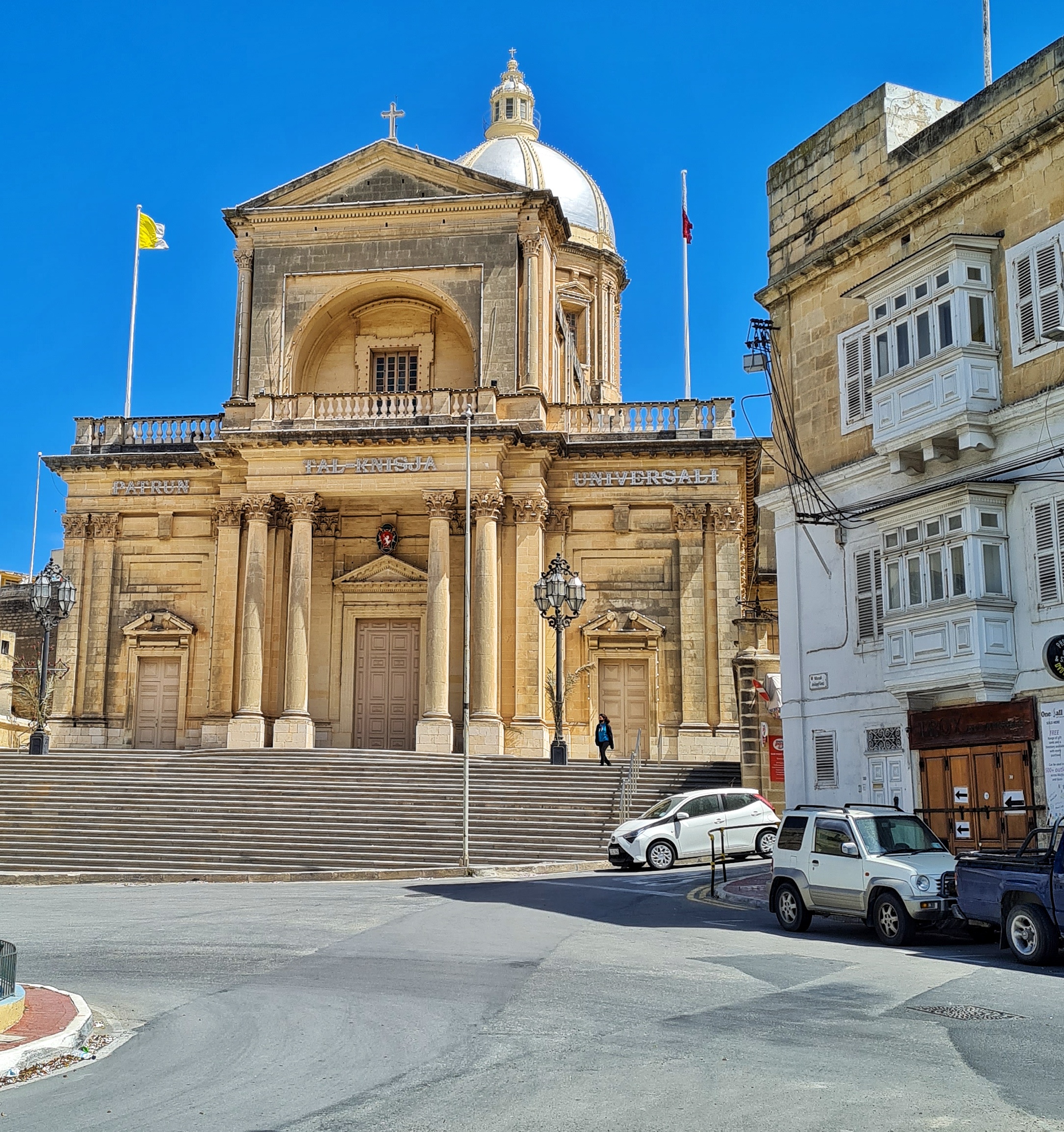 Eine beige Kirche an einer Straße. Rechts ein weiteres Gebäude und parkende Autos. Zur Kirche führen mehrere niedrige Treppenstufen empor.  Die drei Türen (die mittlere mit Säulen davor) sind geschlossen. Darüber eine Dachterrasse mit einem Aufbau in der Mitte. Dort weht eine gelb-weiße Flagge. Im Hintergrund ein runder Turm mit einem kuppelförmigen Dach und einem Türmchen darauf.