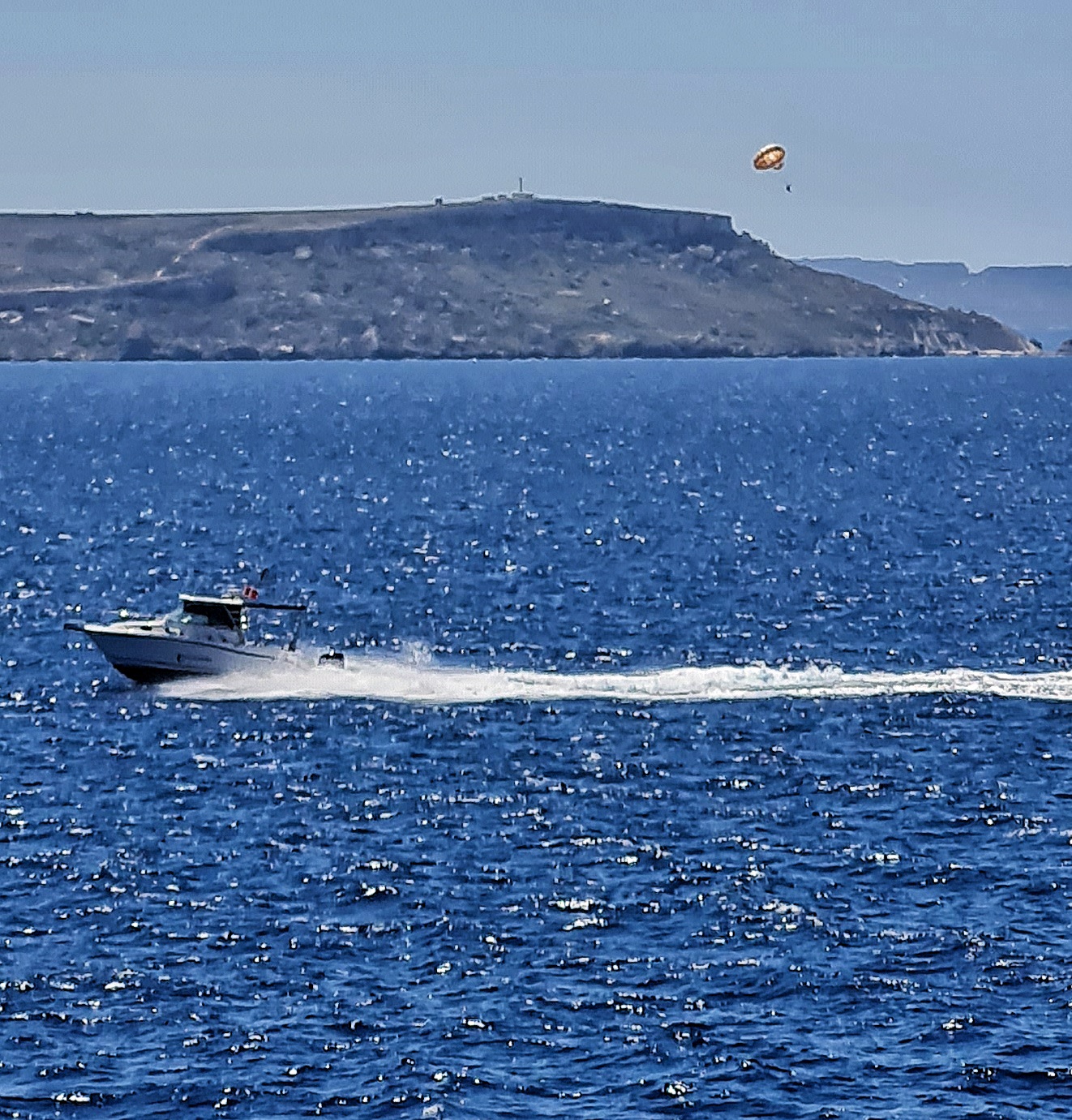Tiefblaues Meer, auf dessen Wellen das Wasser glitzert. Von rechts nach links fährt ein Boot mit einer langen weißen Heckwelle. Im Hintergrund eine ansteigende Landzunge. Darüber schwebt ein Mensch an einem Schirm.
