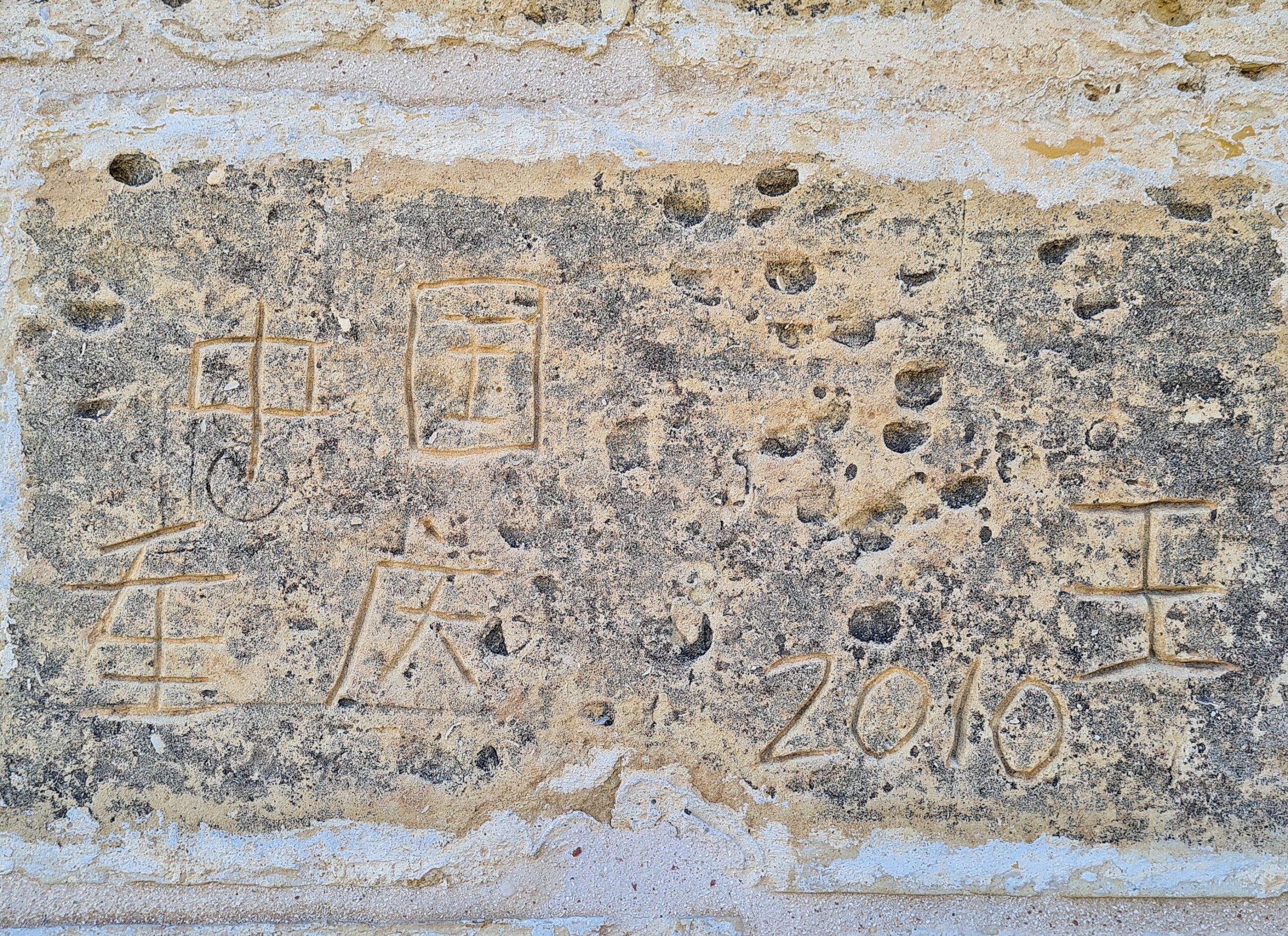 Ein ziemlich abgeblätterter Stein, in den Schriftzeichen (chinesisch oder ähnliches) und 2010 eingeritzt sind.