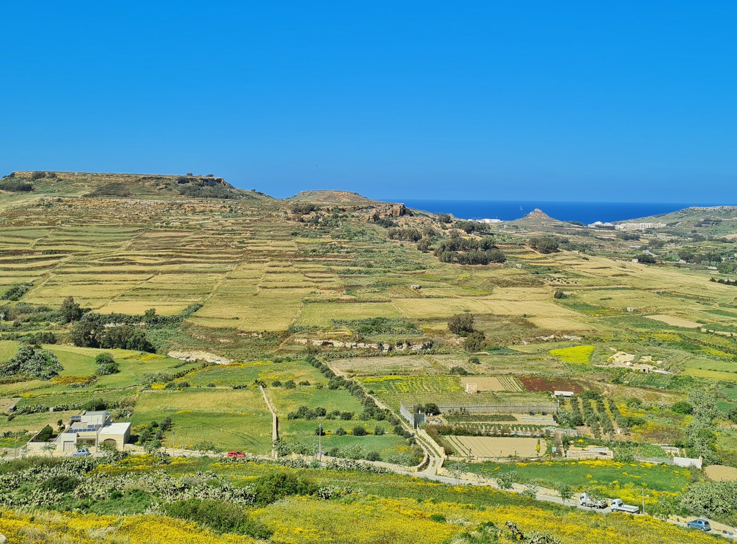 Blick von oben auf eine leicht grüne Ebene mit einzelnen Baumgruppen. Im Hintergrund blaues Meer. Auf der Fläche unten eine Straße und ein Weg, der von ihr abgeht, einzelne Gebäude und Fahrzeuge.