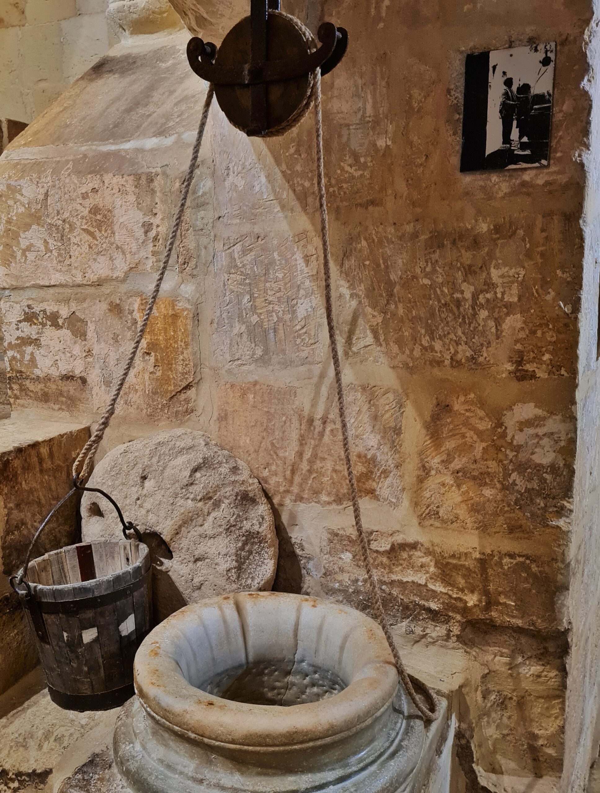 Eine helle Mauer aus Steinen im Inneren. Oben hängt eine große Seilwinde. Die beiden Seile führen nach unten zu einem Holzeimer und einem großen Steingefäß, das oben eine vasenartige Form hat. Es ist nach oben offen. An der Wand hinter dem Holzeimer lehnt eine runde Steinplatte. Oben rechts an der Wand ist ein Schwarz-Weiß-Foto, auf dem Menschen zu sehen sind.in