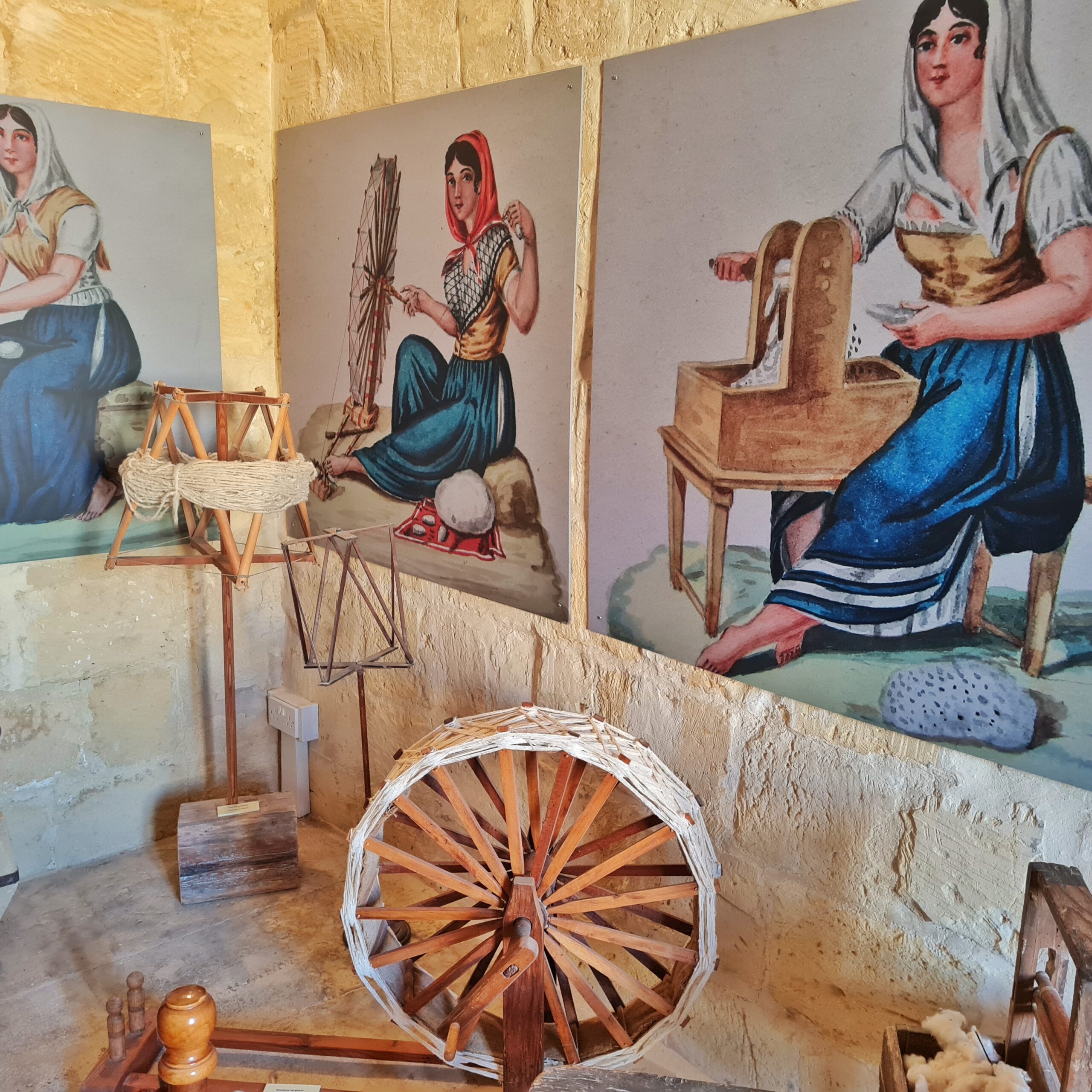 An der Wand Zeichnungen von drei jungen Frauen un dirndlartiger Tracht mit weißen oder roten Kopftüchern. Sie arbeiten an Maschinen aus Holz.. Davor stehen Gerätschaften aus Holz. An einem aus mehreren Holzstäben gebildeten Ständer hängt ein Seilknäuel. Außerdem ist ein große Holzrad zu sehen (das wohl Teil eier Maschine ist). Es besteht außen aus weißem Flechtwerk.Im Inneren führen an beiden -seiten des Flechtwerks mehrere Holzstreben zur Nabe des Rades. Die Abstände zwischen ihnen sind gering.
