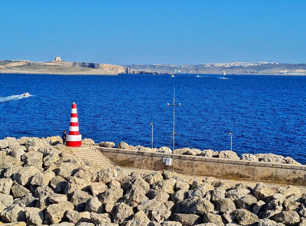 Im Vordergrund eine Kaimauer, davor sind zum Wasser hin, Steine verteilt. Der Weg hinter der Kaimauer führt über Stufen zu einem kegelförmigen Turm, der rot-weiß gestreift ist. Daneben steht eine Frau. Der Turm ist etwa dreimal so groß wie sie. Neben dem Weg sind weitere Steine aufgetürmt. Hinter der Kaimauer blaues Meer, auf dem einige weiße Schnellboote fahren. Links hinten ist eine Landzunge mit Felsen und einem kleinen viereckigen Turm. Ganz hinten ist ebenfalls Land zu sehen, wobei man auf einem Hügel Bebauung erkennen kann.