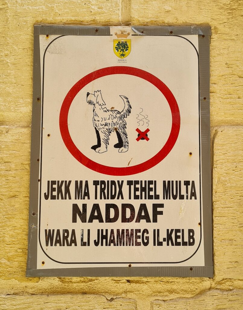 Ein weißes Schild. Oben ist ein Hund mit einem dampfenden Hundehaufen gezeichnet. Darum ein roter Kreis. Auf dem Hundehaufen ist ein rotes X gezeichnet. Darunter die Worte Jekk Ma Tridx Tehel Multa Naddaf Wara Li Jhammeg Il-Kelb.