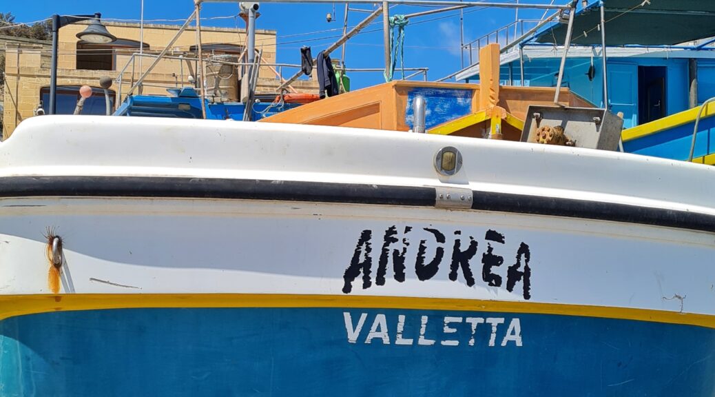Das Heck eines breiten Bootes, das an Land liegt. Im Hintergrund sind Gebäude zu sehen. Das Boot hat unten einen schmalen dunkelroten Streifen, darüber einen breiten blauen und ganz oben ist es weiß. Auf dem Heck steht: Andrea Valletta