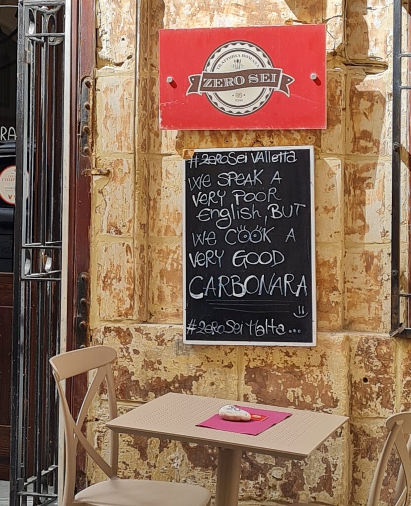 Außen vor einem kleinen Restaurant. Links ist ein Teil der Eingangstür zu sehen. Unten im Bild ein einfacher Tisch mit zwei Stühlen, darauf eine Serviette, die beschwert wird durch einen flachen Stein mit "20" darauf. Darüber an der Hauswand hängt eine schwarze Tafel, auf der mit weißer Kreide geschreben steht: #ZEROSEI VALLETTA WE SPEAK A VERY POOR ENGLISH, BUT WE COOK A VERY GOOD CARBONARA: Über der Tafel hängt ein rotes Schild mit der Aufschrift: ZEROSEI TRATTORIA ROMANA.