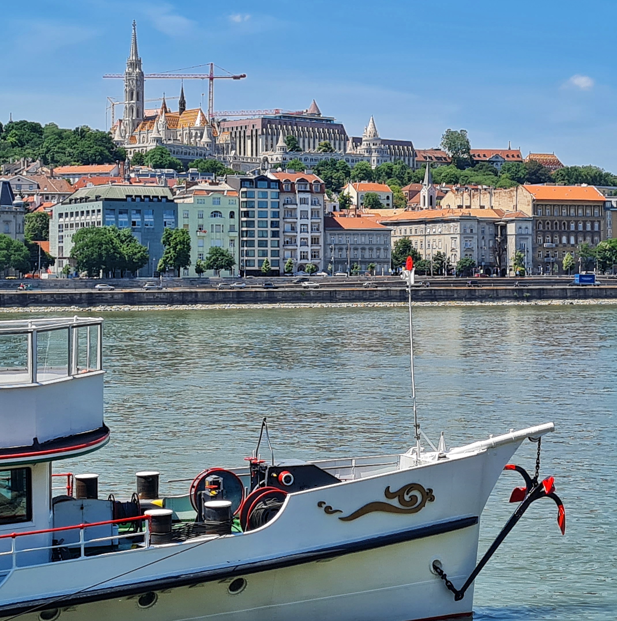 Der Vorderteil eines weißen Schiffes im Vordergrund. Dahinter ein breiter Fluß. Auf der anderen Uferseite mehrere dicht an dicht stehende hohe Gebäude. Darüber eine große Kirche und ein hohes Gebäude.