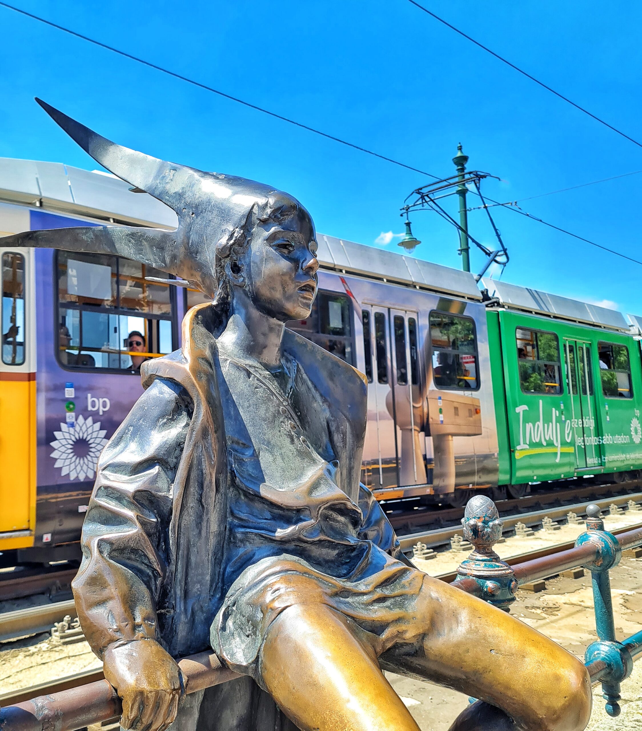 Eine Statue. Im Hintergrund fährt eine Straßenbahn vorbei. Die Statue zeigt ein auf dem Geländer sitzendes größeres Kind in Hosen mit einer geöffneten Jacke. Auf dem Kopf hat es eine Kopfbedeckung mit langen Zipfeln nach hinten.