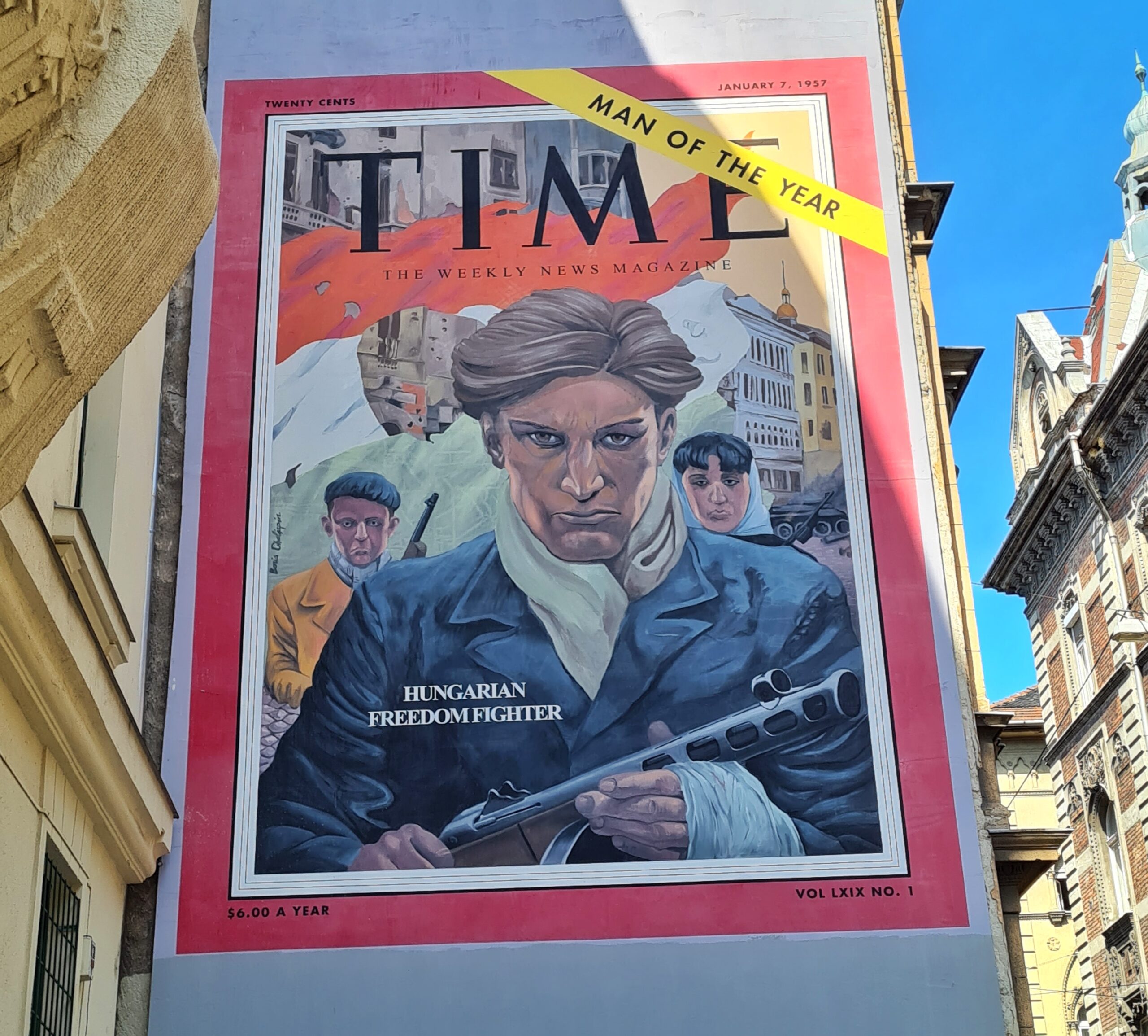 An einer weißen Hauswand das große gemalte Titelblatt der "Time" mit roter Umrandung. Darauf ein Mann mit einem Maschinengewehr in der Hand. Hinter ihm zwei weitere Personen mit Gewehren. Dahinter Gebäude, ein Panzer und weiße Fahnen. Ganz oben steht in kleiner Schrift links "Twenty Cents" und rechts "January 7, 1957". Darunter groß "TIME" und darunter in kleiner Schrift "The Weekly News Magazine". Oben rechts quer über die Ecke ein gelber Balken mit schwarzer Schrift "Man of the Year". Und auf der Zeichnung des Mannes mit dem Maschinengewehr steht "Hungarian Freedom Fighter".
