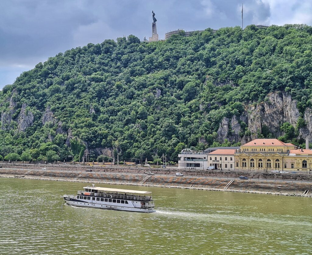 Ein Hügel mit Felsen und Bäumen und Büschen. Oben ist ein festungsartiges Gebäude mt einer Fahnenstange zu sehen. Daneben auf einem hohen Steinturm eine große Metallfigur. Vor dem Hügel fließt ein Fluß, auf dem ein Ausflugsschiff von rechts nach links fährt. Am Fuß des Hügels ist ein langgestrecktes Gebäude.